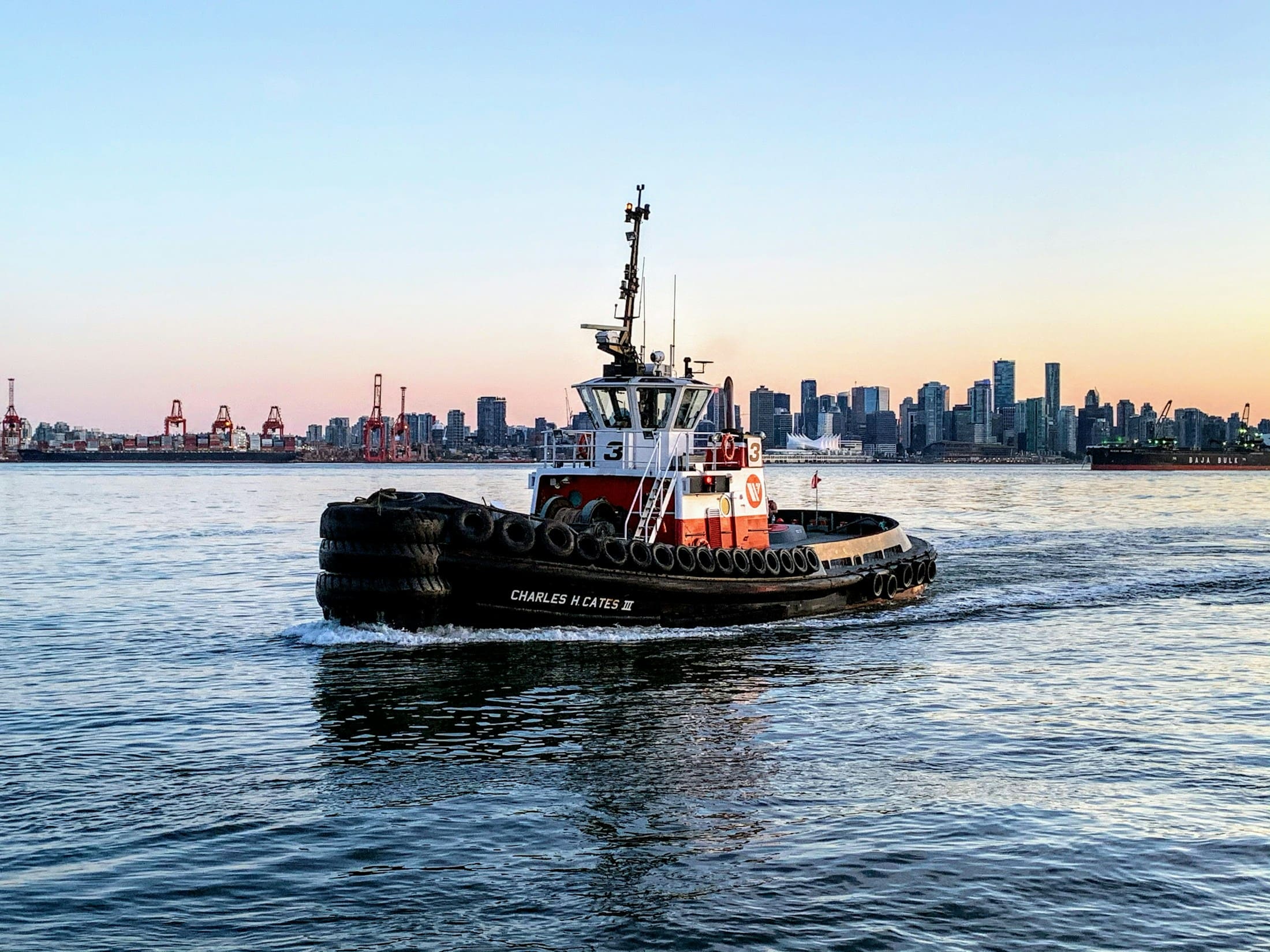A tugboat on calm harbour water at dusk, a city skyline glowing behind it.
