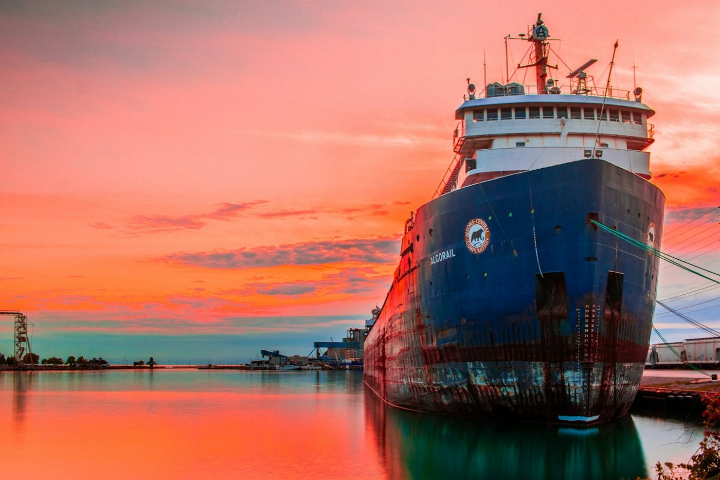 A large ship moored at a dock beneath a vivid coral-red sunset sky.